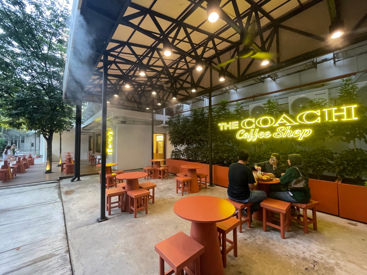 На изображении кафе "The Coachi Coffee Shop" с двумя людьми за столом, сидящими лицом друг к другу. Кафе расположено в помещении с высоким потолком, покрытым светлым полотном, и открытым пространством с