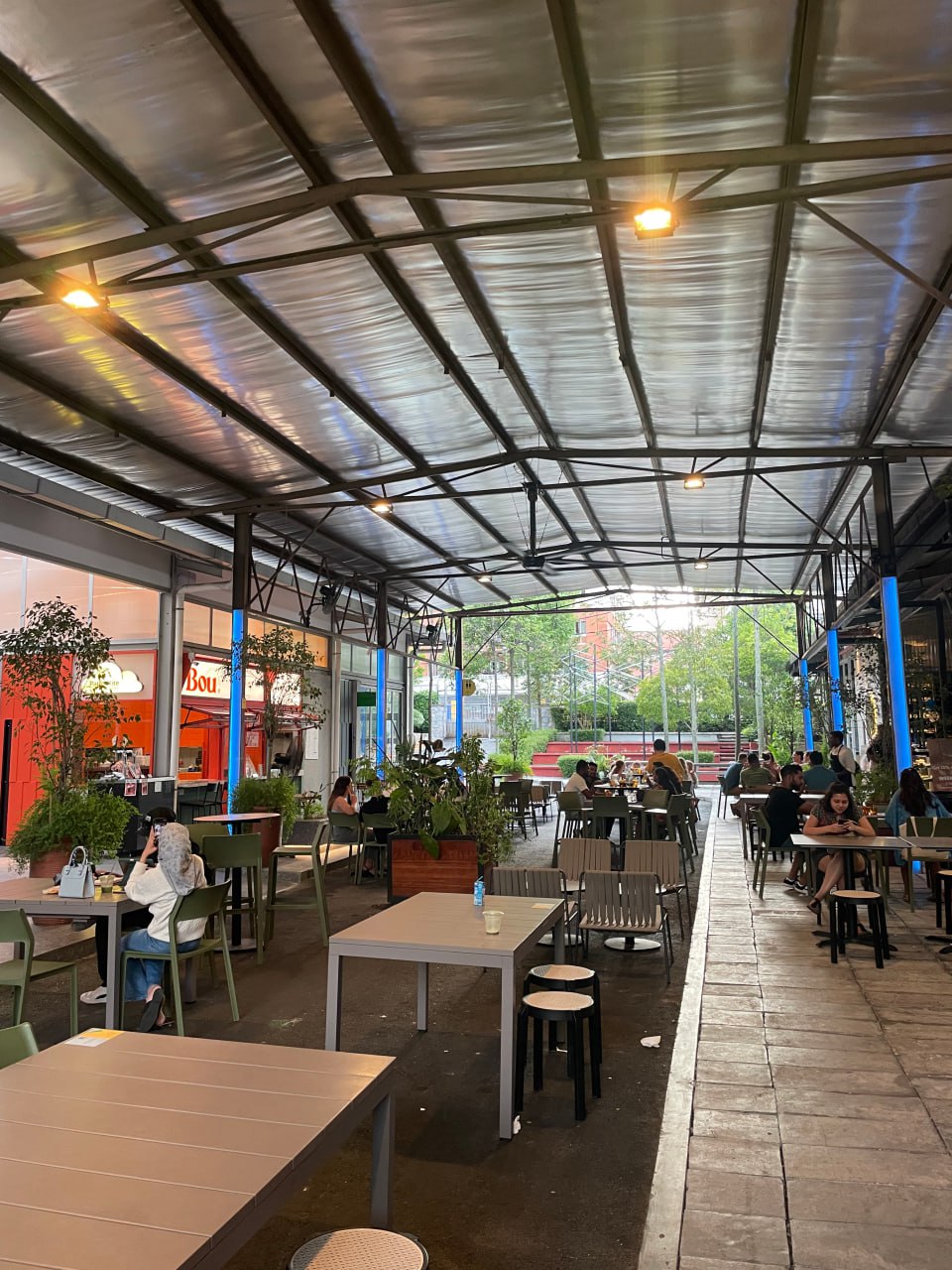 Здесь изображен открытый внутренний дворик кафе, где сидят несколько человек за столами, а также несколько растений и столы. В центре изображения видна длинная стена, за которой расположены окна кафе, а также несколько деревьев и зданий на