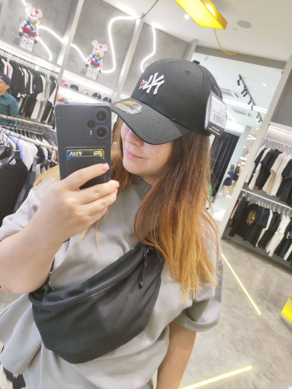 Фотография женщины, которая держит в руке смартфон, снимая себя в зеркале, в магазине одежды. На ней капюшон с логотипом New York Yankees, чехол для телефона, который закрывает заднюю часть телефона,
