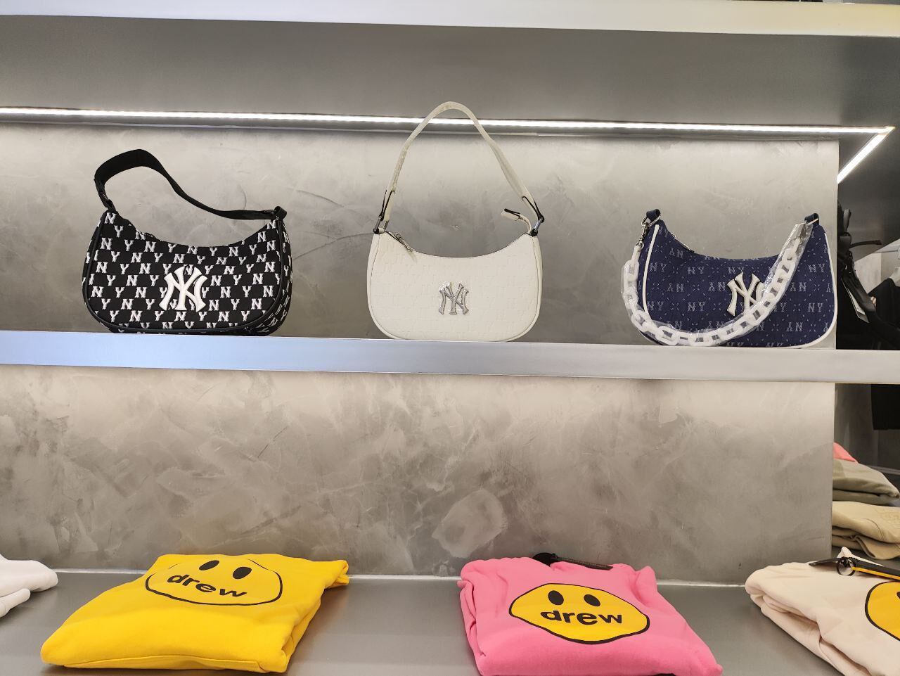 Представлены три женские сумки, расположенные на полке, и три женских футболки с изображением улыбающегося лица, расположенные на полке ниже. Сумки расположены на полке сверху, а футболки - на полке