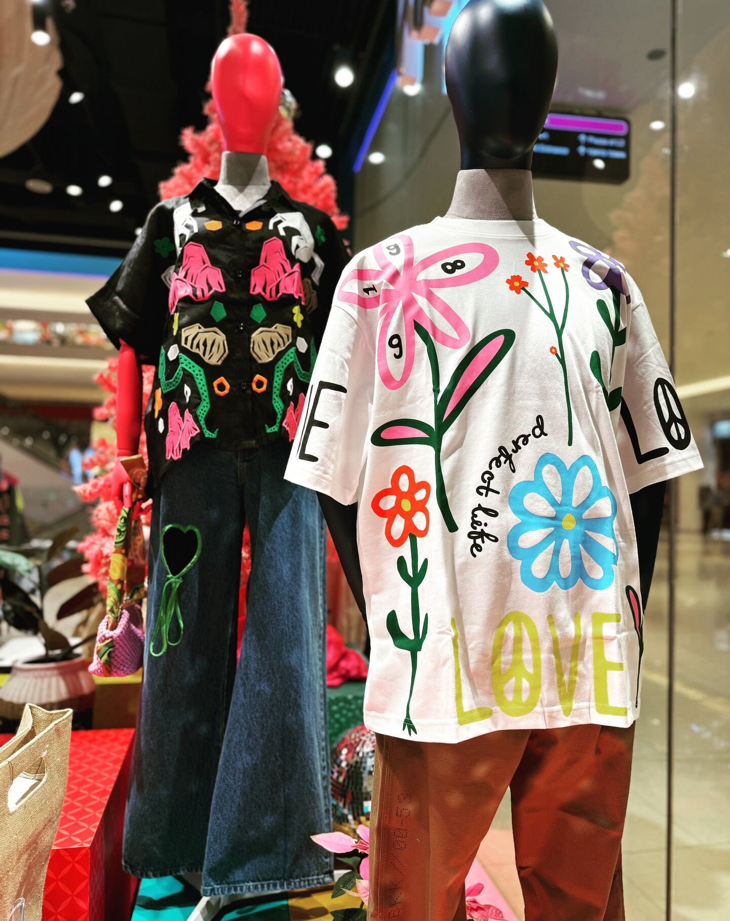 В этом изображении две манекены в витрине магазина, стоящие рядом друг с другом. Манекен слева надет на чёрную рубашку с цветочным узором и синими джинсами. Манекен справа надет на белую рубаш