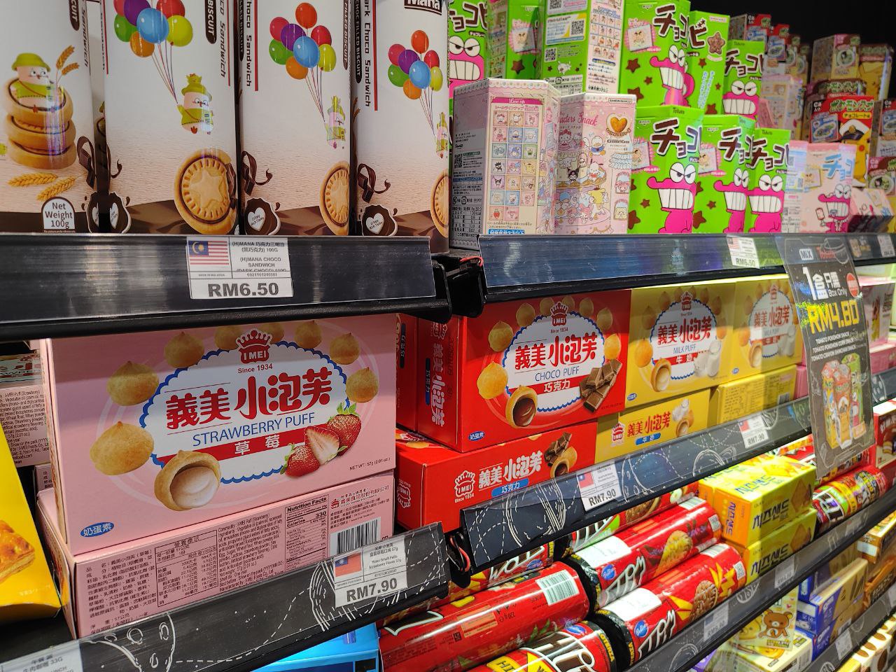 Это изображение полки с продуктами в магазине, на которой расположены коробки с пирожными, конфетами, чипсами и другими продуктами. На полке есть несколько коробок с пирожными, в том числе одна с изображением на коробке, которая