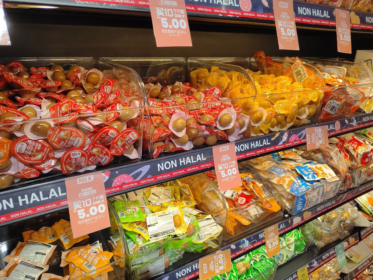 На изображении полки с различными продуктами, в том числе снеками и сладостями, которые продаются в магазине. На полках есть яркие ярлыки с надписью "Non Halal", что означает, что продукты не соответствуют мусульманским