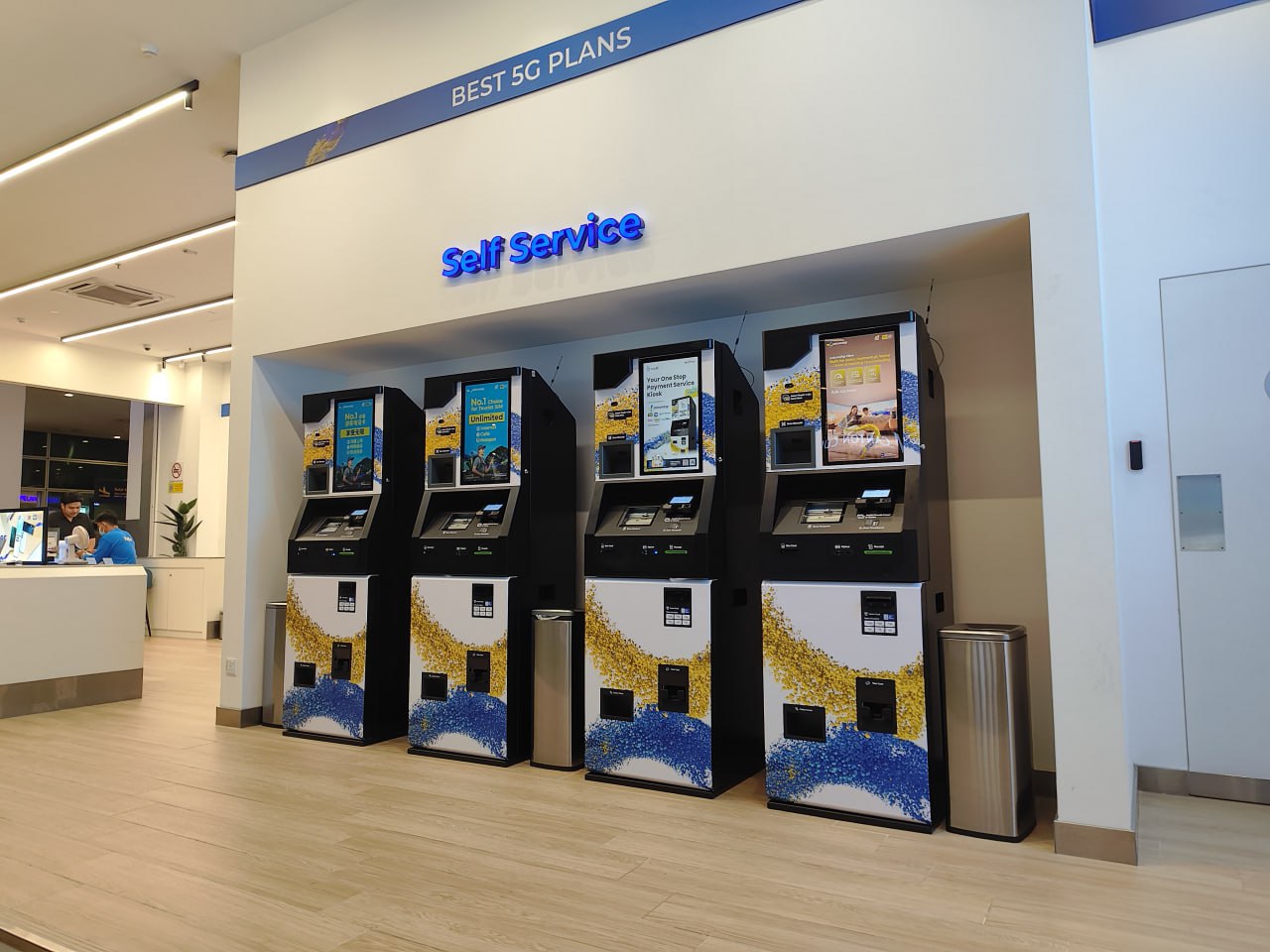 Это изображение внутреннего пространства магазина, где на стене выставлены четыре автомата для самообслуживания, над которыми написано "BEST 50 PLANS" и "Self Service". В центре стены расположен небольшой вход в помещение, а слева от