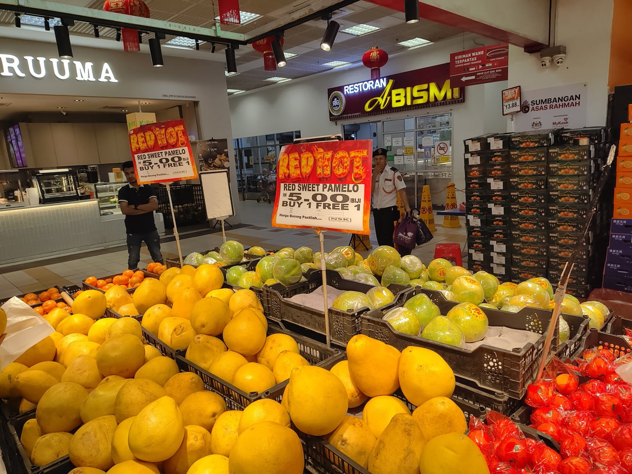На изображении показан магазин с большим количеством фруктов, в том числе апельсинов, гранатов и мандаринов, а также с двумя плакатами с текстом "RED HOT RED SWEET PAMLO BUY 1 FREE 1".