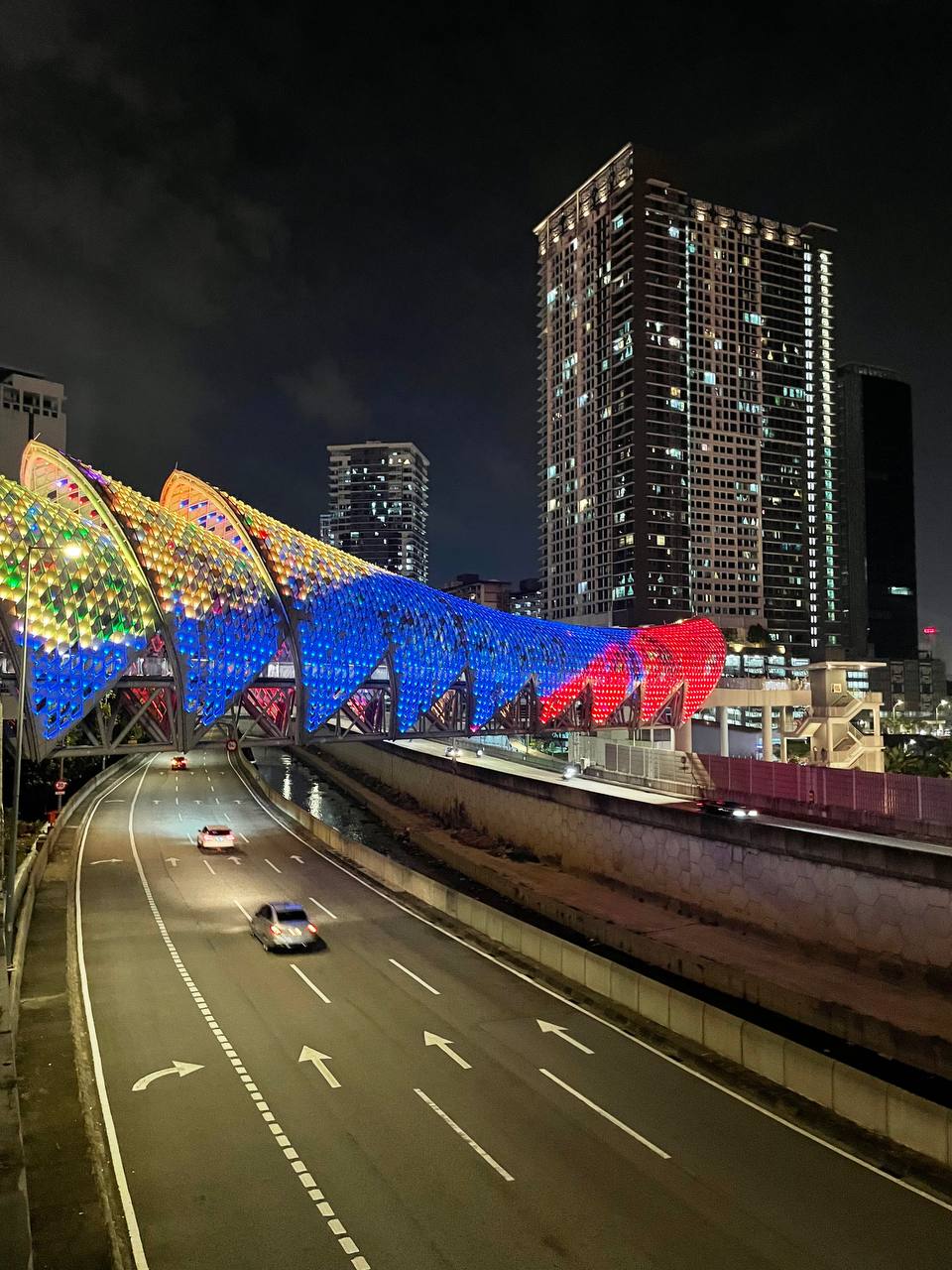 На изображении ночной городской пейзаж, включающий мост, автомобильный тоннель, несколько высоких зданий и автомобиль, движущийся по дороге.