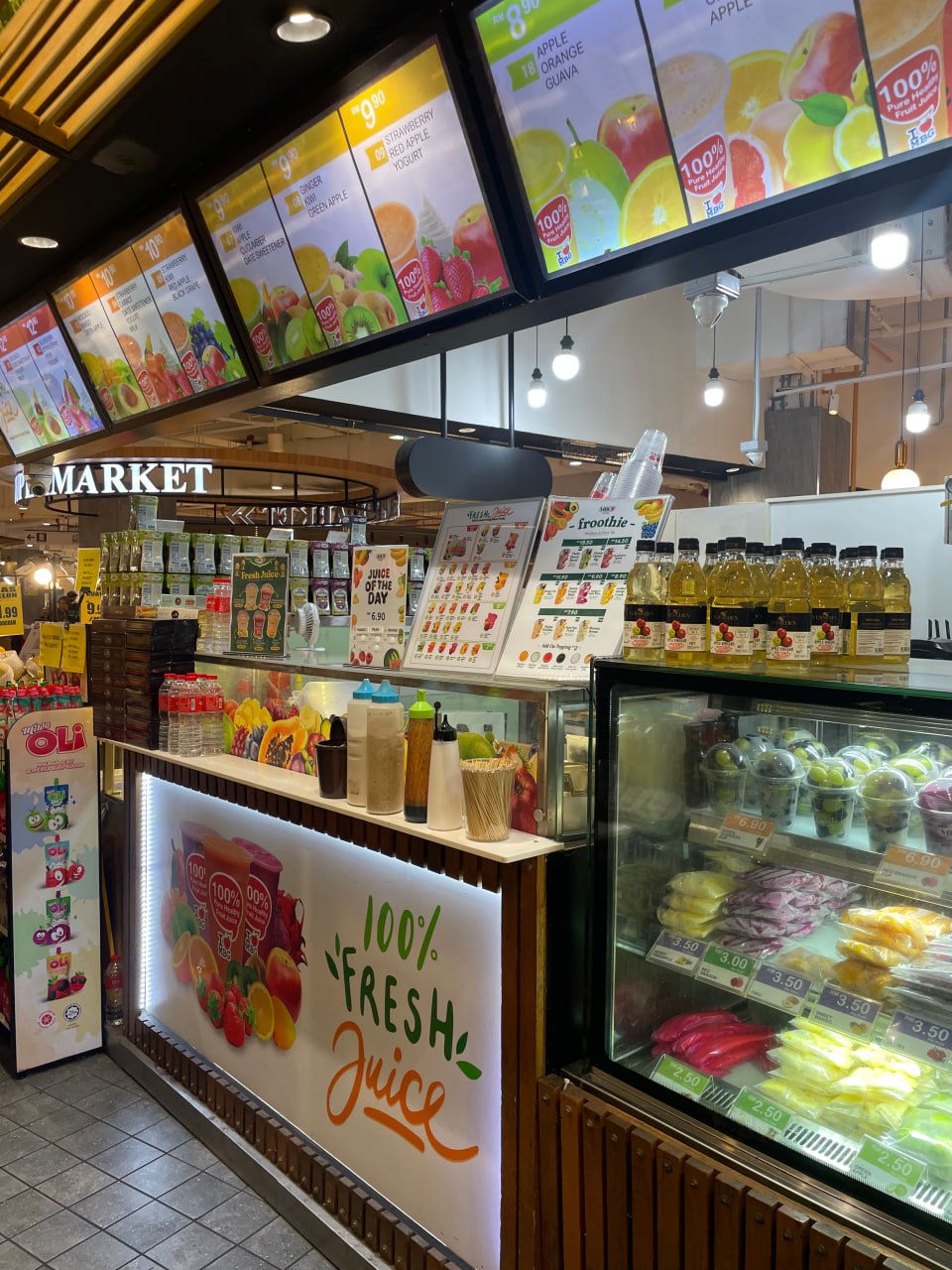Магазин с фруктовыми напитками и фруктами. В центре изображения - бар с напитками, справа - холодильник с фруктами. На баре написано "100% FRESH Juice". На потолке - рекламные плакаты с изображением фруктов