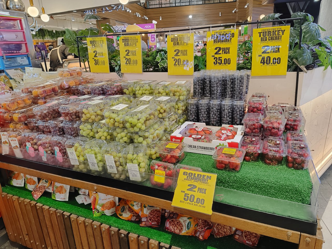 Это изображение торгового павильона, где на витрине продается фрукты и овощи. На витрине есть яркие ярлыки с ценами, а также зеленая искусственная трава. На витрине также есть несколько коробок с фруктами, а также несколько