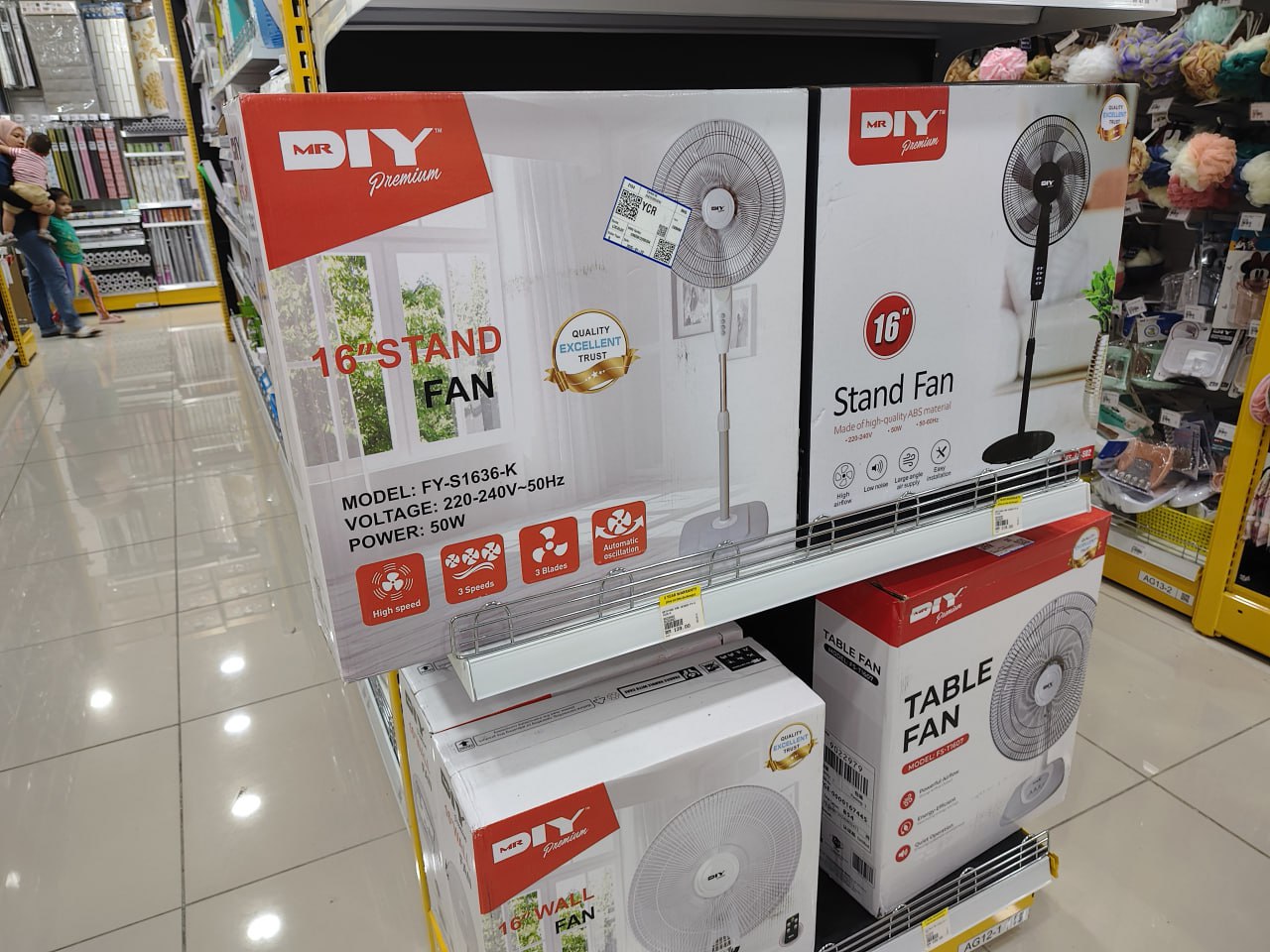 На этом изображении полка с товарами в магазине, на которой расположены коробки с электрическими фанов, стойки для фанов и коробки с таблицевыми фановами. На коробках написано "MR DIY" и "DIY". На