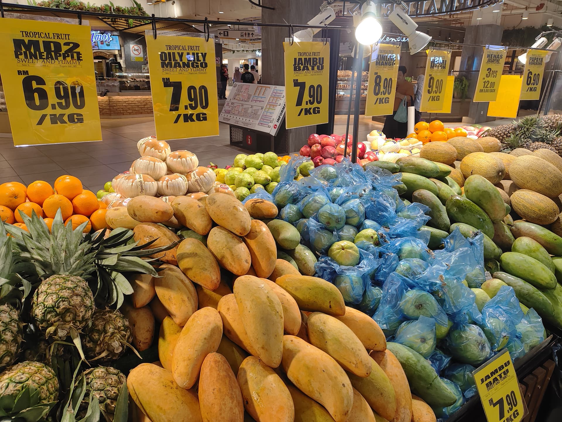 На изображении продавец фруктов в супермаркете. В продаже различные фрукты, в том числе мандарины, апельсины, манго, ананасы, яблоки, персики, гранаты, груши, дыни,